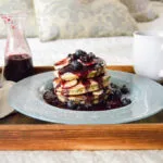 A close-up of a spoon dripping thick Blueberry Balsamic Syrup onto a stack of golden-brown pancakes, highlighting its deep purple color.