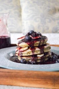 A beautifully styled brunch setup featuring a plate of Banana Bread Pancakes, a jar of homemade Blueberry Balsamic Syrup, and a cup of coffee on a rustic wooden table.