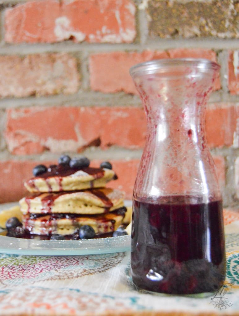 A close-up of a small pitcher pouring deep purple Blueberry Balsamic Syrup over a warm stack of pancakes, highlighting its rich color and smooth texture.