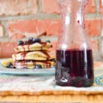 A spoon drizzling thick, glossy Blueberry Balsamic Syrup into a glass jar, surrounded by fresh blueberries on a rustic wooden table.