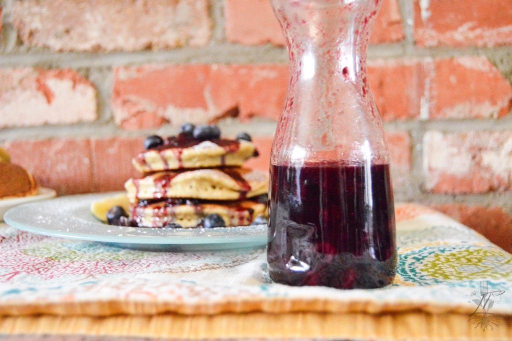 A spoon drizzling thick, glossy Blueberry Balsamic Syrup into a glass jar, surrounded by fresh blueberries on a rustic wooden table.