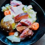 Close-up of ham shank simmering in a large pot surrounded by fresh vegetables.