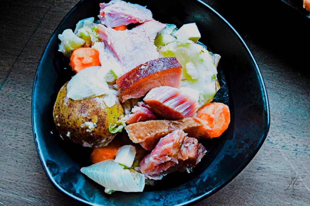 Close-up of ham shank simmering in a large pot surrounded by fresh vegetables.