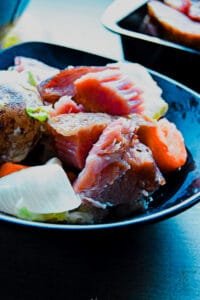 Close-up of ham shank simmering in a large pot surrounded by fresh vegetables.