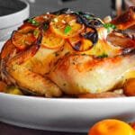 Golden-brown roasted chicken topped with charred lemon slices, served on a white platter with roasted potatoes and fresh herbs.