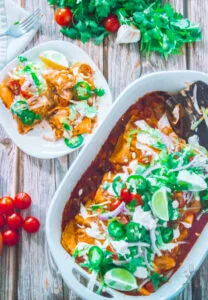Plate of served beef enchiladas next to a baking dish, garnished with cilantro, tomatoes, garlic, and lime.