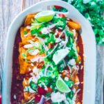 Overhead view of a white baking dish filled with beef enchiladas topped with sour cream, sliced jalapeños, onions, tomatoes, and fresh lime wedges.