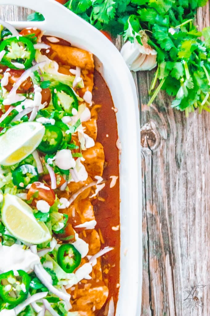 Overhead close-up of beef enchiladas in a white baking dish, topped with vibrant fresh garnishes like jalapeños, lime wedges, cherry tomatoes, red onion, and a drizzle of sour cream, with garlic and cilantro on a rustic wooden table.