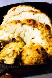 Front view of a whole roasted cauliflower sliced open to reveal its soft interior, topped with a browned garlic parmesan coating.