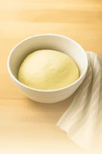 A smooth ball of pizza dough rising in a white ceramic bowl with a striped towel on the side, set on a light wood surface.