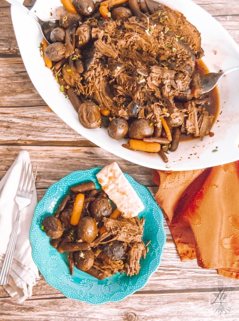 Overhead view of shredded roast beef with carrots and potatoes in a white serving dish and plated on a turquoise plate with a cracker.