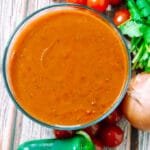 Bowl of rich, red enchilada sauce surrounded by fresh jalapeños, tomatoes, garlic, onion, and cilantro on a rustic wooden table.