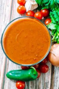 Bowl of rich, red enchilada sauce surrounded by fresh jalapeños, tomatoes, garlic, onion, and cilantro on a rustic wooden table.