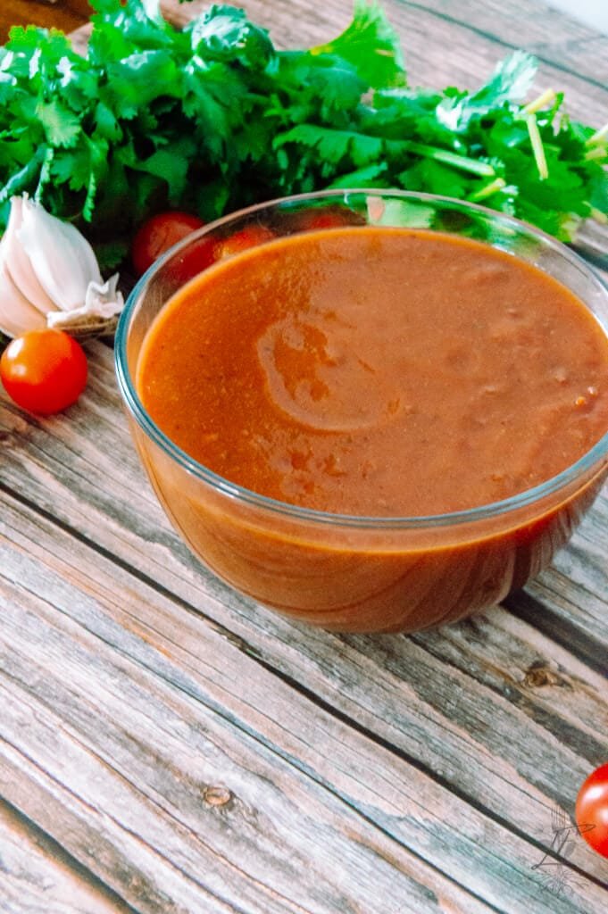 Side view of a bowl filled with chipotle enchilada sauce, surrounded by tomatoes, garlic, and fresh herbs.