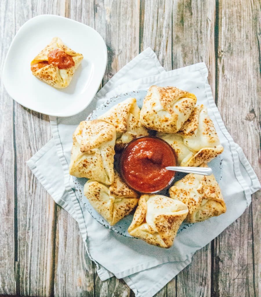Top-down view of a serving platter filled with baked pizza pockets and a dish of marinara, with one pocket plated separately.