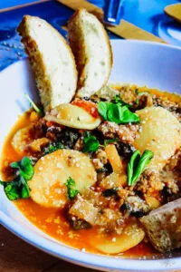 Close-up of sausage and pepper ravioli in a white bowl topped with basil and parmesan, served with crusty bread.