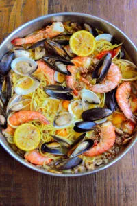 Top-down view of a full skillet of seafood pasta with shrimp, mussels, clams, grilled lemons, and spaghetti on a rustic wood table.