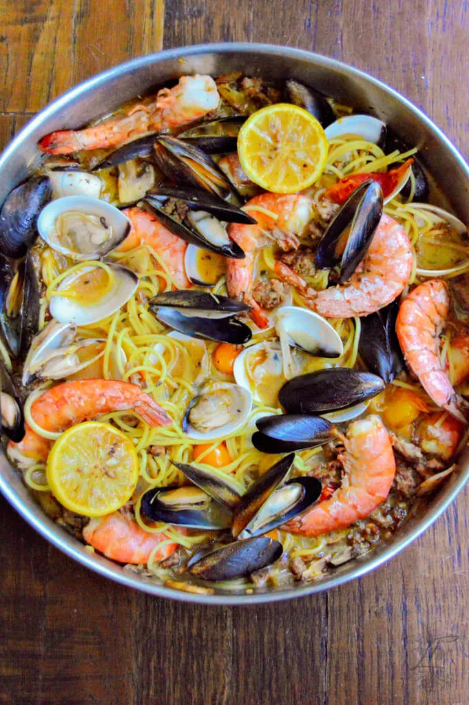 Top-down view of a full skillet of seafood pasta with shrimp, mussels, clams, grilled lemons, and spaghetti on a rustic wood table.