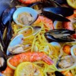 Side view of a pan filled with shrimp, clams, mussels, spaghetti, and grilled lemons in a golden broth.