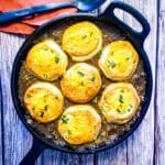 Golden brown biscuits baked on top of savory beef pot pie filling in a cast iron skillet, garnished with herbs.