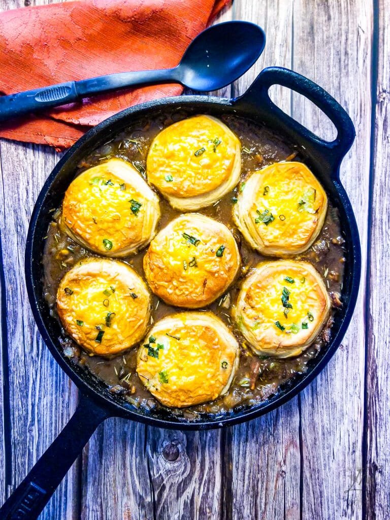 Golden brown biscuits baked on top of savory beef pot pie filling in a cast iron skillet, garnished with herbs.