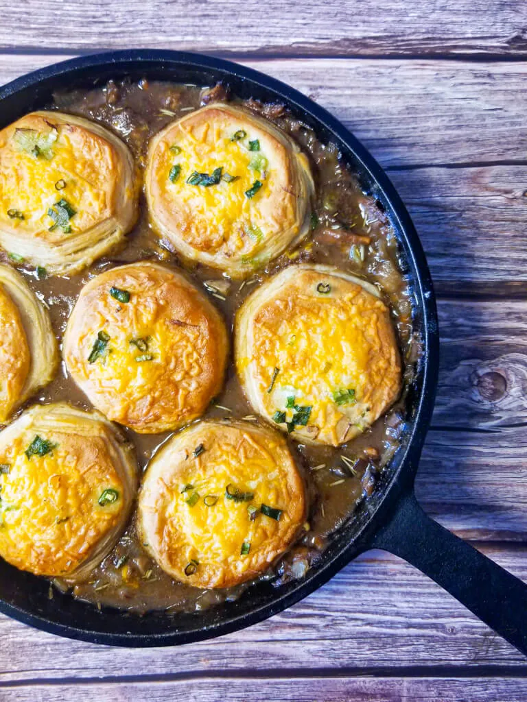 Close-up of cheesy biscuit-topped beef pot pie in a cast iron skillet with thick, flavorful gravy beneath.