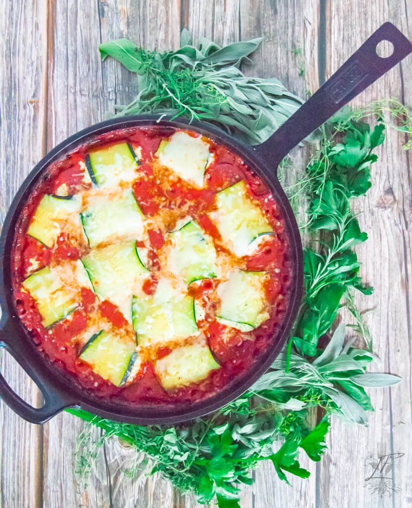 Overhead view of a full cast iron skillet filled with baked zucchini rollatini, surrounded by fresh herbs.