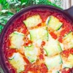 Cheesy baked zucchini rollatini in a cast iron skillet surrounded by fresh herbs on a rustic wood table.