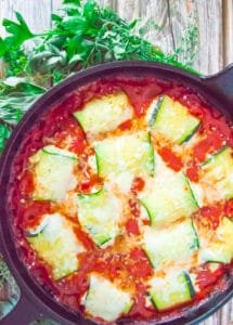 Cheesy baked zucchini rollatini in a cast iron skillet surrounded by fresh herbs on a rustic wood table.