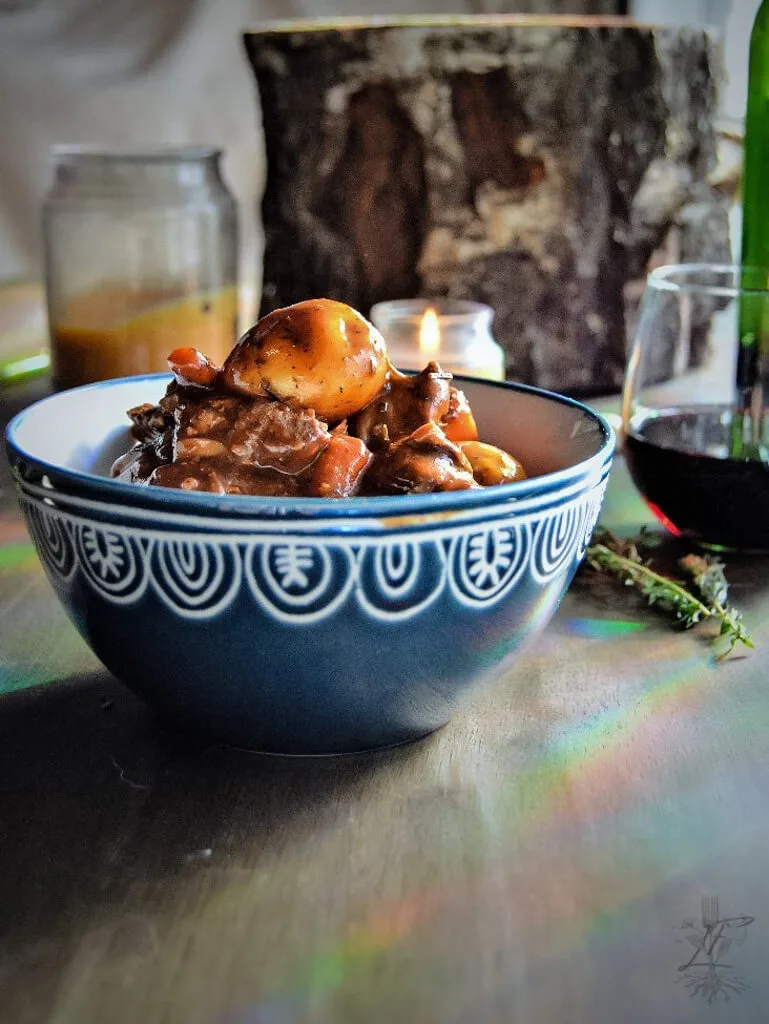 Side view of a bowl of beef stew on a wood table with herbs, candles, and a glass of red wine in the background.
