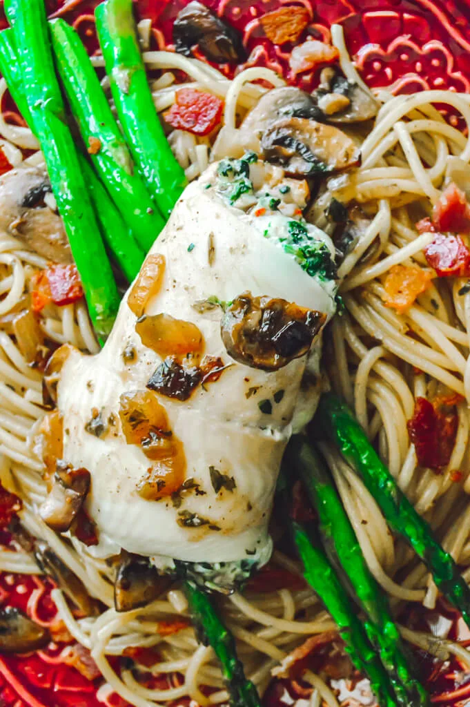 Overhead shot of stuffed flounder fillet served over mushroom bacon pasta with asparagus spears on a red plate.