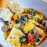 Side angle of a bowl of ravioli with crumbled sausage, summer squash, cherry tomatoes, and fresh herbs, served with garlic bread.