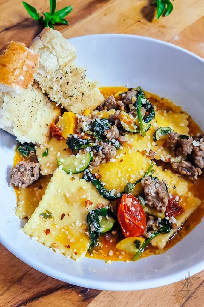 Side angle of a bowl of ravioli with crumbled sausage, summer squash, cherry tomatoes, and fresh herbs, served with garlic bread.