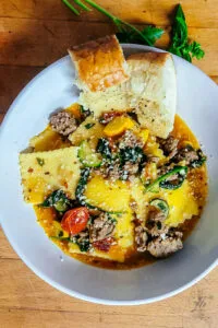 Overhead view of a white bowl filled with ravioli, ground sausage, zucchini, tomatoes, spinach, and garlic bread on a wooden surface.