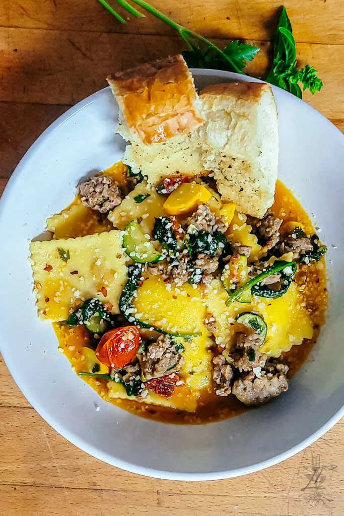 Overhead view of a white bowl filled with ravioli, ground sausage, zucchini, tomatoes, spinach, and garlic bread on a wooden surface.