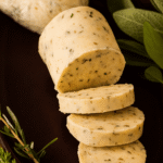 Autumn herb butter log with sage and seasonal herbs, sliced into rounds on a dark surface