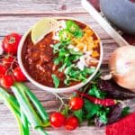 Bowl of chili with cheese, sour cream, jalapeños, cilantro, and lime wedge, surrounded by fresh vegetables.