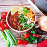 Bowl of chili with cheese, sour cream, jalapeños, cilantro, and lime wedge, surrounded by fresh vegetables.