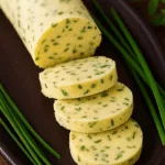 Sliced chive compound butter log on dark plate with fresh chive.