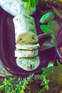 Sliced herb butter log on dark surface with rosemary, thyme, and sage.