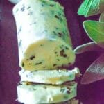 Poultry herb butter log with sage, thyme, and rosemary, sliced into rounds on a dark surface.